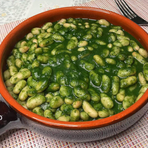 Fagioli cannellini in una salsa verde di spinaci e cipolla rossa, speziati con sale, pepe, coriandolo e origano. Serviti in un contenitore di terracotta.