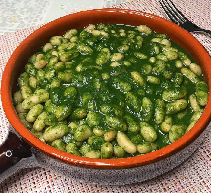 Fagioli cannellini in una salsa verde di spinaci e cipolla rossa, speziati con sale, pepe, coriandolo e origano. Serviti in un contenitore di terracotta.